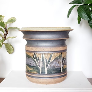May include: A cylindrical ceramic vase with a landscape design. The vase has a dark grey body with a beige rim and base. The landscape design includes white birch trees, green and purple hills, and a beige background.