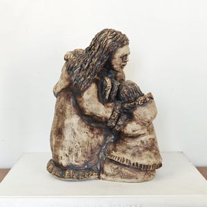 May include: A tan and dark brown ceramic sculpture depicting two figures in an embrace. The larger figure has long textured hair and wears a hooded garment with a fringed hem, while the smaller figure is positioned in front, also wearing a hooded coat.