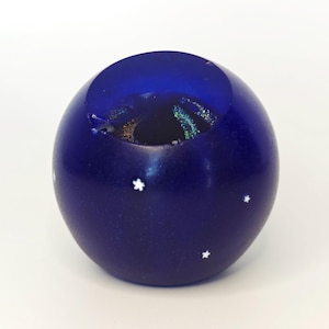 May include: A dark blue, spherical glass object with a hollowed-out top. The interior reveals iridescent colours. Three small white star-shaped designs are evenly spaced on the surface. The object is likely a decorative piece.