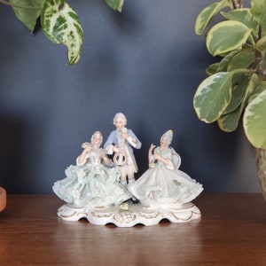 May include: A porcelain figurine set of three people, two women and one man, dressed in vintage clothing. The women are wearing light green dresses with lace details, and the man is wearing a blue jacket and white pants. The figurines are standing on a white base with gold accents.