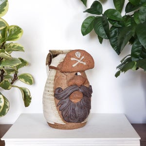 Peut inclure: Pichet en céramique sur le thème des pirates, avec un chapeau, une barbe et une moustache bruns. Le pichet a un corps blanc et marron avec une anse. Le chapeau de pirate présente un motif de tête de mort et d'os croisés.