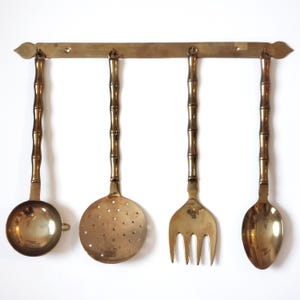 Peut inclure: Ensemble de quatre ustensiles de cuisine dorés avec des manches de style bambou suspendus à un support mural. Les ustensiles comprennent une louche, une cuillère à fentes, une fourchette et une cuillère.