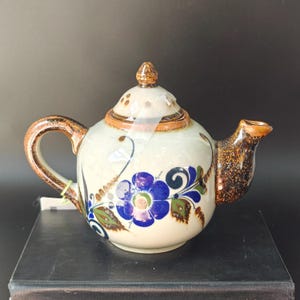 May include: A round ceramic teapot with a cream-coloured base, featuring hand-painted blue flowers and green leaves. The handle, spout, and lid finial have a speckled brown glaze. A clear adhesive strip is visible across the lid and body.