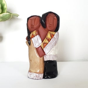 May include: A ceramic sculpture of two figures embracing. The figures are painted in shades of brown, white, and yellow. The sculpture is a representation of a couple in love.