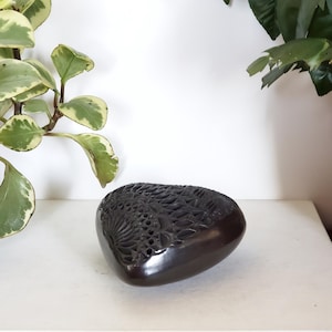 May include: A dark brown, heart-shaped decorative object with detailed carved floral patterns. The object has a smooth, glossy finish and is placed on a white surface. Green leafy plants are in the background.