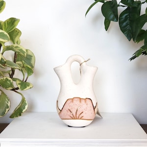 Peut inclure: Vase double en céramique blanche avec un motif rose et or. Le vase a une forme unique avec une poignée reliant les deux ouvertures.
