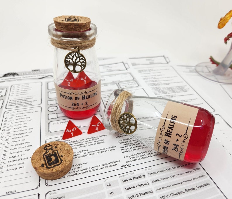 May include: Two glass potion bottles with cork stoppers and labels that read "Potion of Healing 2D4+2". The bottles are filled with a red liquid and have a small tree charm hanging from the top. Two red dice are in front of the bottles.