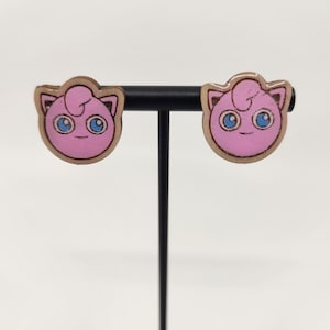May include: A pair of pink and blue Jigglypuff earrings. Each earring features a pink Jigglypuff face with blue eyes and a brown outline. The earrings are displayed on a black stand against a white background.