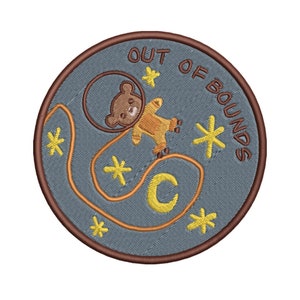 May include: A round, blue embroidered patch with a brown border. The patch features a cartoon bear wearing a space suit and roller skates, a crescent moon, and stars. The text "OUT OF BOUNDS" is written around the edge of the patch.