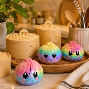 May include: Three round, rainbow-colored dumpling figurines with large black eyes and pink cheeks, arranged alongside two light-colored bamboo steamer baskets on a round wooden tray. The dumplings feature a smooth, glossy finish.