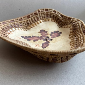 May include: Handwoven basket with a natural, organic aesthetic. The basket features a light-colored, central panel adorned with a dark brown floral design. The rim and sides are crafted from woven, light brown reeds, creating a rustic, textured appearance.