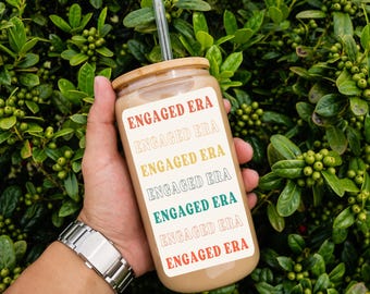 May include: A brown reusable tumbler with a bamboo lid and a clear straw. The tumbler has the text "ENGAGED ERA" repeated in different colors.