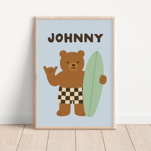Puede incluir: Impresión enmarcada con un oso pardo sosteniendo una tabla de surf. El oso lleva pantalones cortos a cuadros y hace el gesto shaka. El nombre "JOHNNY" está impreso encima del oso sobre un fondo azul claro. El marco es de color madera clara.