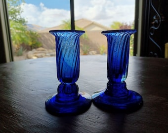May include: Two cobalt blue glass candle holders with a swirl design. The candle holders have a wide base and a narrow top.