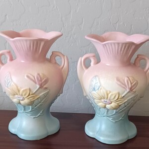 May include: Two ceramic vases with a floral design. The vases are in a pastel color scheme, with a pink top, cream middle, and light blue base. Each vase has handles and a flower and leaf motif.