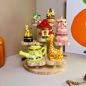 May include: A wooden tiered display holds several decorative trinket boxes. The boxes are shaped like a giraffe, a watering can, a mushroom, a carousel, and other whimsical designs. The boxes are brightly colored with intricate details.