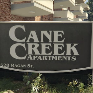 May include: A sign for Cane Creek Apartments, with white lettering on a dark gray background. The sign also displays the address 1528 Ragan St. The sign is mounted on white posts and is surrounded by grass and foliage.
