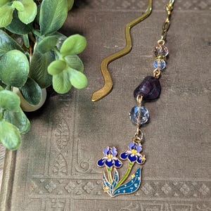 May include: A gold-toned metal bookmark with a wavy design, featuring a dangling charm chain. The chain includes clear faceted beads, a dark purple tumbled stone, and a gold-rimmed enamel charm depicting two blue iris flowers with green stems.