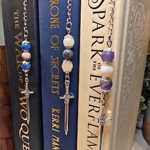May include: Three beaded bookmarks with silver sword charms hang from the top of books. The bookmarks feature blue, white, and gray beads, and purple and white beads. The books have titles like "The War of Two Queens" and "Spark of the Everflame."