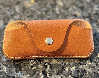May include: A brown leather glasses case with a white stitch detail and a silver button closure.