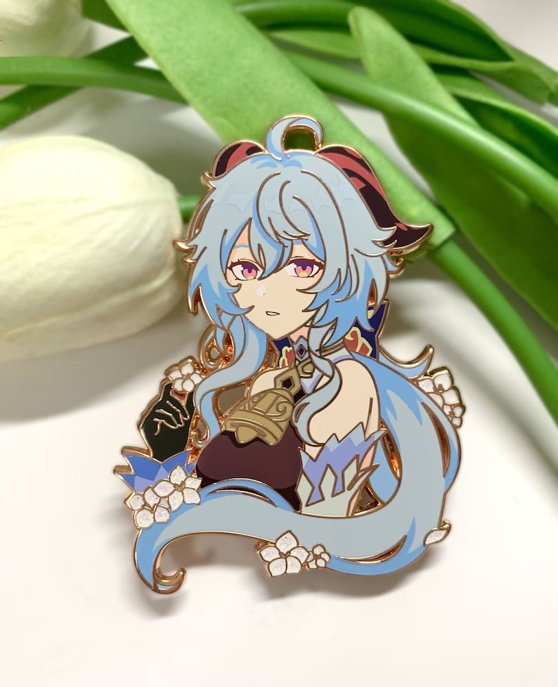 Op de afbeelding: Een gouden emaille pin met een cartoonfiguur met lang blauw haar, gekleed in een paars-blauwe outfit met een bel. Het personage is omringd door witte bloemen.