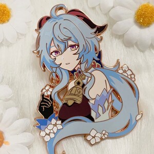 Op de afbeelding: Een gouden emaille pin met een personage met lang blauw haar, een rood-zwart hoofdband en een gouden bel. Het personage draagt een wit-blauwe outfit met een rood-gouden ontwerp. De pin is omringd door witte bloemen.
