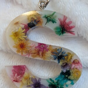 May include: A clear resin letter "S" keychain filled with colorful dried flowers. The flowers include yellow, pink, purple, and blue blooms. A silver chain is attached to the top of the letter.