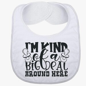 Puede incluir: Babero blanco para bebé con el texto negro "I'M KIND of a BIG DEAL AROUND HERE". El babero tiene forma redondeada con un bolsillo y dos almohadillas absorbentes blancas en la parte superior. Dos gráficos de chupete a cada lado del texto.
