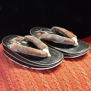 May include: A pair of black wooden geta sandals with brown straps and floral designs. The sandals are on a red knitted surface.
