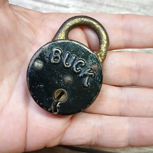 May include: A vintage, round padlock with the word "BUCK" embossed on its black face. The lock has a keyhole and a gold-colored shackle. The metal shows signs of age and wear, giving it a rustic appearance.