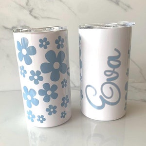 May include: Two white stainless steel tumblers with lids. One tumbler has a blue floral pattern and the other has the name "Eva" written in blue cursive.