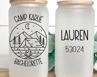 Puede incluir: Dos vasos de vidrio esmerilado con tapas de bambú y pajitas. El vaso de la izquierda tiene una ilustración en blanco y negro de un campamento con montañas, una tienda de campaña, una fogata y estrellas. El texto "Camp Karlie Bachelorette" está impreso en el vaso. El vaso de la derecha tiene el texto "Lauren 5.30.24" impreso en él.