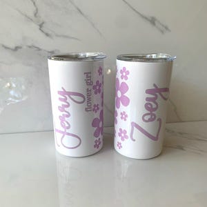May include: Two white stainless steel tumblers with pink floral designs and personalized names. The tumbler on the left says "Jerry flower girl" and the tumbler on the right says "Zoey".