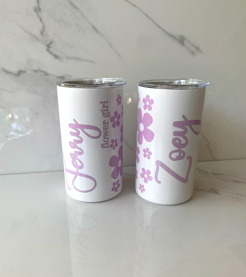 May include: Two white stainless steel tumblers with pink floral designs and personalized names. The tumbler on the left says "Jerry flower girl" and the tumbler on the right says "Zoey".
