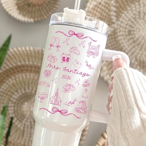 May include: White insulated tumbler with a clear lid and handle. The tumbler is decorated with pink line art of wedding-themed illustrations, including champagne glasses, rings, and a wedding cake. The text "Mrs. Santiago 2026" is also featured.
