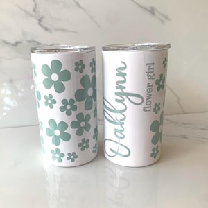May include: Two white stainless steel tumblers with lids. One tumbler has a pattern of green flowers and the other has the name "Kallynn" and the text "flower girl" in gray.