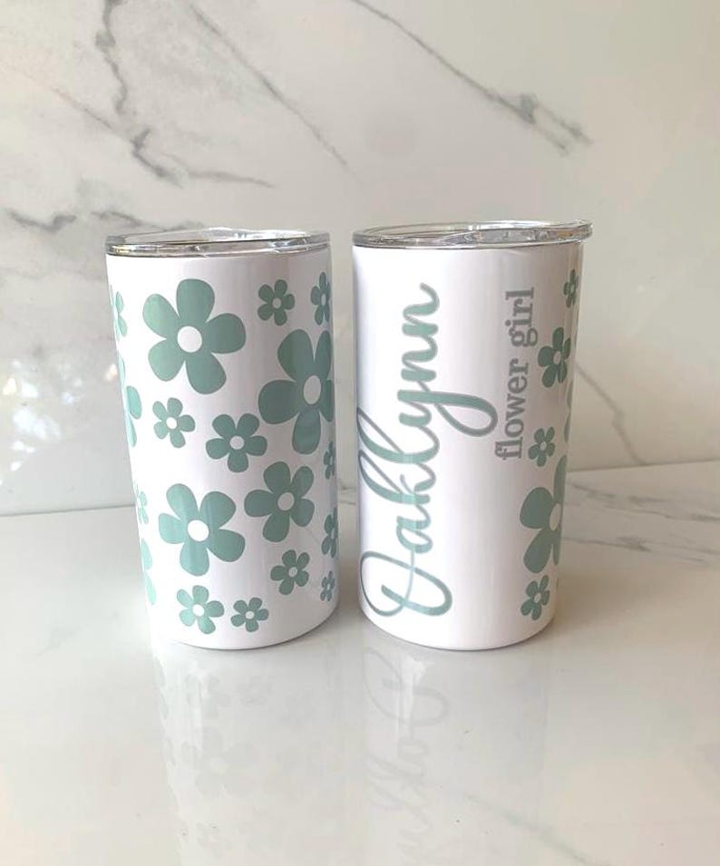 May include: Two white stainless steel tumblers with lids. One tumbler has a pattern of green flowers and the other has the name "Kallynn" and the text "flower girl" in gray.