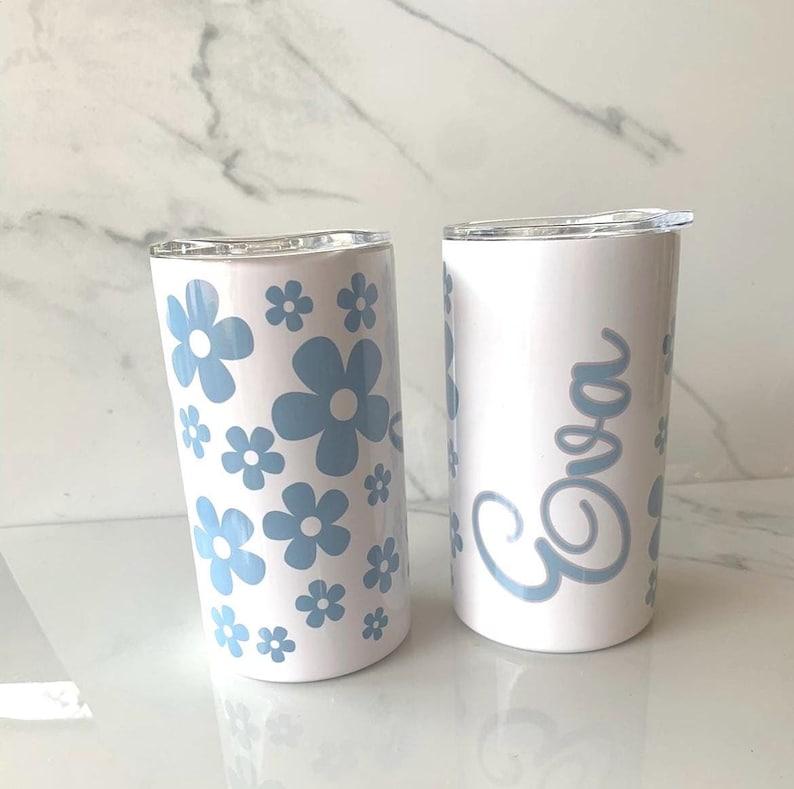 May include: Two white stainless steel tumblers with lids. One tumbler has a blue floral pattern and the other has the name "Eva" written in blue cursive.