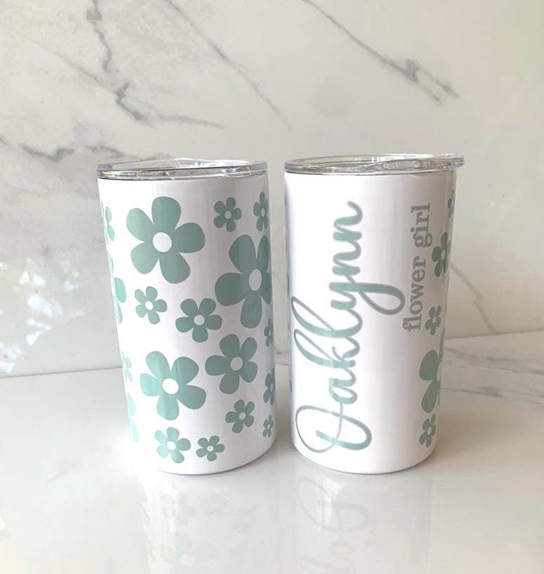 May include: Two white stainless steel tumblers with lids. One tumbler has a floral pattern with green flowers. The other tumbler has the name "Oaklynn" and the text "Flower girl" written in cursive.