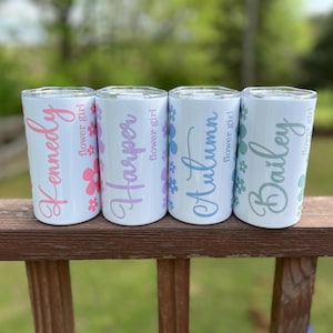 May include: Four white stainless steel tumblers with personalized names and flower girl text. The tumblers are arranged in a row on a wooden railing. The names are Kennedy, Harper, Autumn, and Bailey.