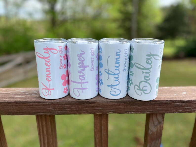 May include: Four white stainless steel tumblers with personalized names and flower girl text. The tumblers are arranged in a row on a wooden railing. The names are Kennedy, Harper, Autumn, and Bailey.