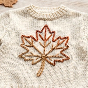 May include: A cream-colored knit sweater featuring a large, embroidered maple leaf design on the front. The leaf is stitched in gradient shades of brown, orange, and gold. The sweater has a ribbed crew neckline and long sleeves, showcasing a textured yarn construction.
