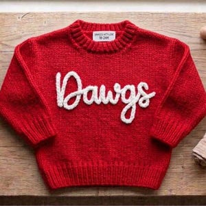 May include: A bright red long-sleeve knit sweater featuring the word Dawgs in white cursive lettering across the chest. The sweater has a ribbed crew neckline, cuffs, and hem. A white interior label displays the text Dawgs With Love 18-24M.
