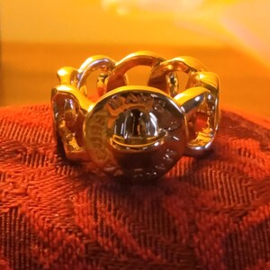 May include: A gold-colored ring with a chain-link design. The ring has a circular centerpiece with embossed details. The ring is displayed on a red textured surface, with a blurred yellow background.