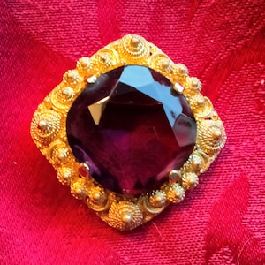 May include: A large, dark purple gemstone set in a gold-toned, ornate, square-shaped frame. The frame is decorated with small, raised circles.
