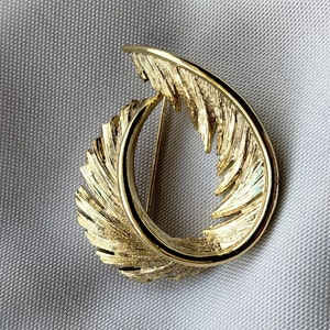 May include: A gold-toned brooch in the shape of a stylized feather. The feather is curled into a circle, creating a unique and elegant design.