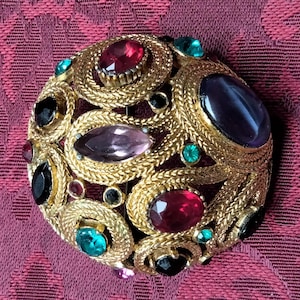 Puede incluir: Un broche dorado con un diseño circular que presenta una variedad de piedras de imitación de colores. El broche está engastado con piedras rojas, rosas, verdes y moradas.