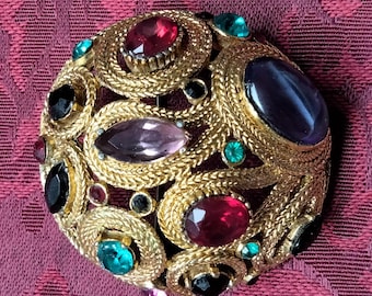 Könnte beinhalten: Eine goldfarbene Brosche mit einem runden Design, das mit verschiedenen bunten Strasssteinen besetzt ist. Die Brosche ist mit roten, rosa, grünen und violetten Steinen besetzt.