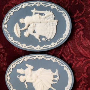 May include: Two oval blue and white jasperware plaques featuring raised relief designs. The top plaque depicts figures with an umbrella and the text APRIL. The bottom plaque shows figures ice skating and the text JANUARY. Both have white borders.