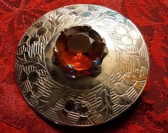 May include: A silver-toned brooch with an engraved floral design. The brooch features a large, faceted, amber-colored gemstone at its center. The brooch is circular and set against a red background.